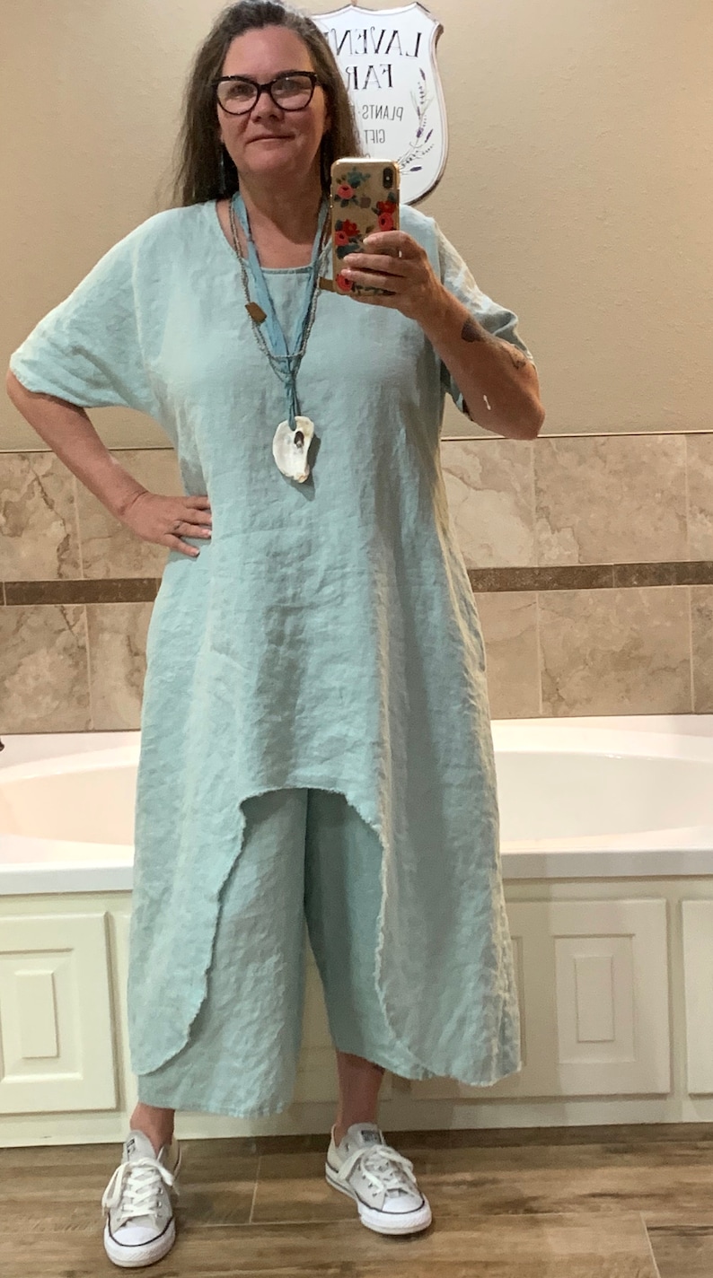 May include: A woman wearing a light blue linen dress with a shell pendant necklace and white sneakers. The dress has a high-low hemline and a relaxed fit. The woman is standing in a bathroom with a white bathtub and tiled walls.