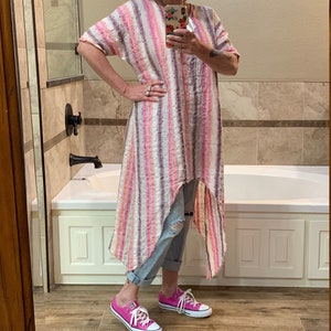 May include: A woman wearing a pink and white striped long dress with a high-low hemline, a brown hat, and pink Converse sneakers. She is standing in a bathroom with a white bathtub.