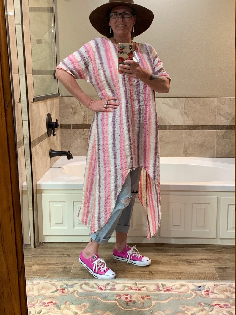 May include: A woman wearing a pink and white striped long dress with a high-low hemline, a brown hat, and pink Converse sneakers. She is standing in a bathroom with a white bathtub.