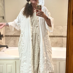 Może przedstawiać: Kobieta w białej, długiej kurtce z koronkowym wykończeniem, noszonej na białej, koronkowej sukience. Ma na sobie czarny cylinder z brązowym piórem i białe buty. Stoi w łazience z białą wanną i białymi kafelkami na ścianach.