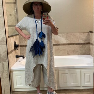 May include: A woman wearing a wide-brimmed straw hat, a linen dress with a blue tassel necklace, and checkered sneakers. She is standing in a bathroom with a white bathtub.