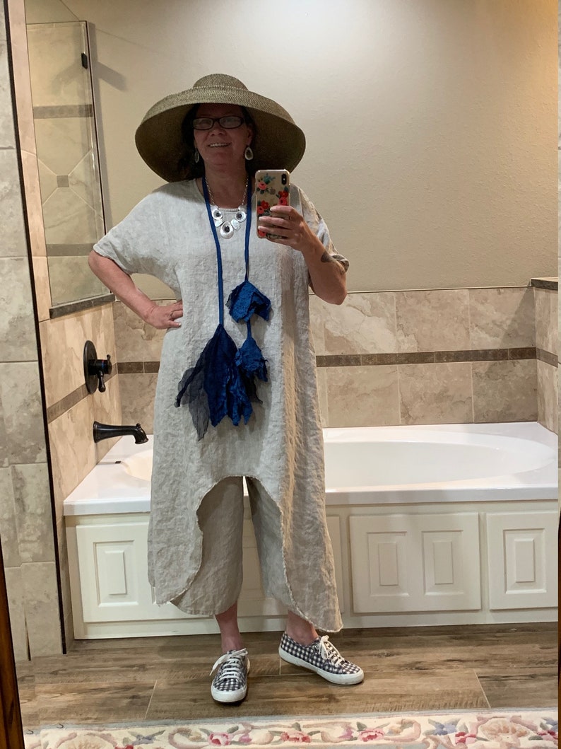 May include: A woman wearing a wide-brimmed straw hat, a linen dress with a blue tassel necklace, and checkered sneakers. She is standing in a bathroom with a white bathtub.