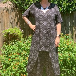 May include: A woman wearing a black and white patterned dress with a black wide-brimmed hat and a silver necklace. She is standing in front of a green bush and a wooden fence.