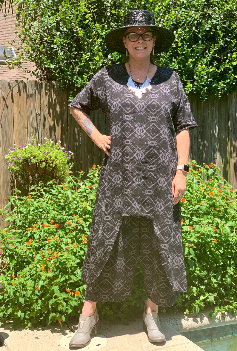 May include: A woman wearing a black and white patterned dress with a black wide-brimmed hat and a silver necklace. She is standing in front of a green bush and a wooden fence.