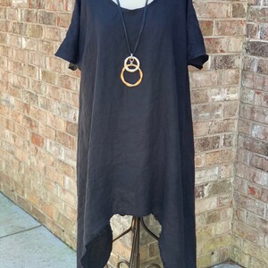 May include: A black linen dress with a high-low hemline and a raw edge finish. The dress has short sleeves and a scoop neckline. A silver and gold chain necklace with a double circle pendant is worn with the dress.