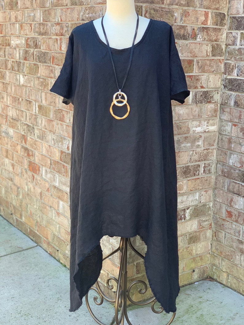 May include: A black linen dress with a high-low hemline and a raw edge finish. The dress has short sleeves and a scoop neckline. A silver and gold chain necklace with a double circle pendant is worn with the dress.