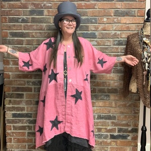 Puede incluir: Una chaqueta de lino rosa con estampado de estrellas negras. La chaqueta tiene cierre de botón y una silueta larga y fluida. La modelo lleva una falda negra y un sombrero de copa negro.