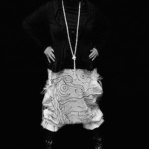 Może przedstawiać: Kobieta w czarnej kurtce, białym naszyjniku z pereł i spódnicy z czarno-białym, wirującym wzorem. Uśmiecha się i nosi okulary.