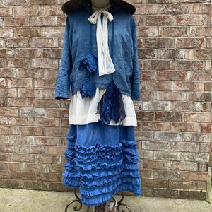 May include: A blue linen dress with a white ribbon and a wide-brimmed straw hat. The dress has a ruffled skirt and a layered top with a white underskirt. The dress is on a mannequin and the mannequin is wearing white boots.