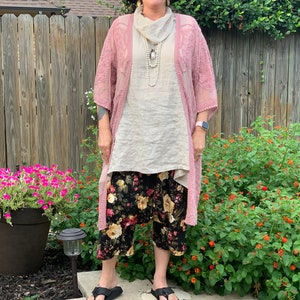 May include: A woman wearing a pink lace cardigan over a white top, black floral print pants, and black sandals. She is wearing a black hat and a pearl necklace.