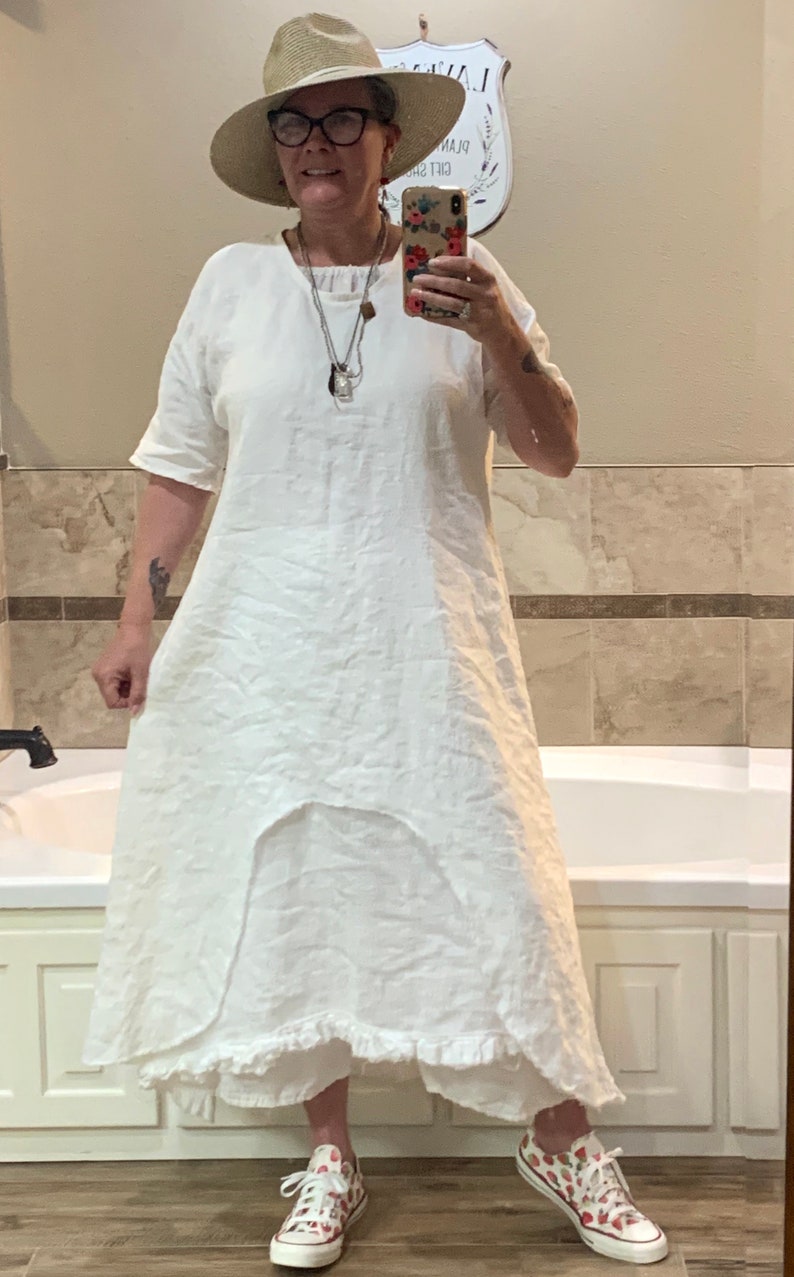 May include: A white linen dress with a ruffled hem and a scoop neck. The dress is worn with a straw hat and strawberry print sneakers.