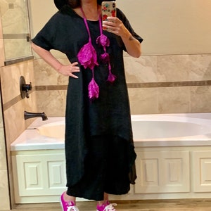 May include: A woman wearing a black maxi dress with a pink flower necklace, a wide-brimmed black hat, and pink sneakers. She is standing in a bathroom with a white bathtub.
