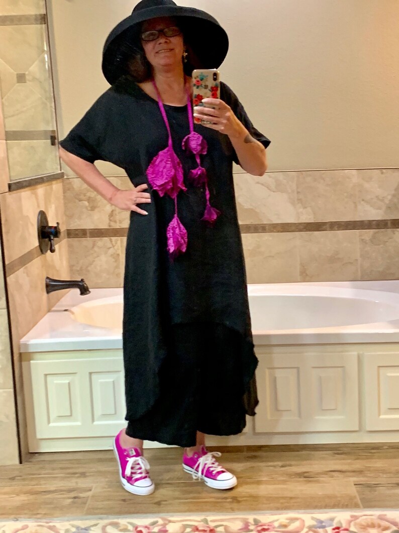 May include: A woman wearing a black maxi dress with a pink flower necklace, a wide-brimmed black hat, and pink sneakers. She is standing in a bathroom with a white bathtub.