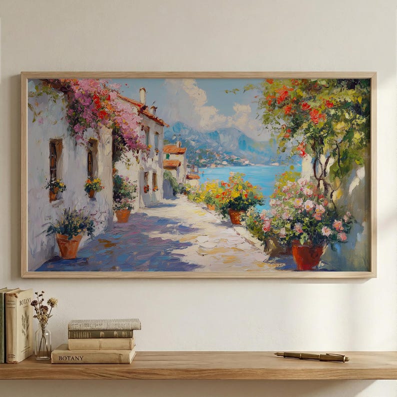 May include: Oil painting depicting a coastal street scene with white buildings, terracotta flower pots, and a view of the blue sea and mountains. The artwork is framed in a light wood frame.