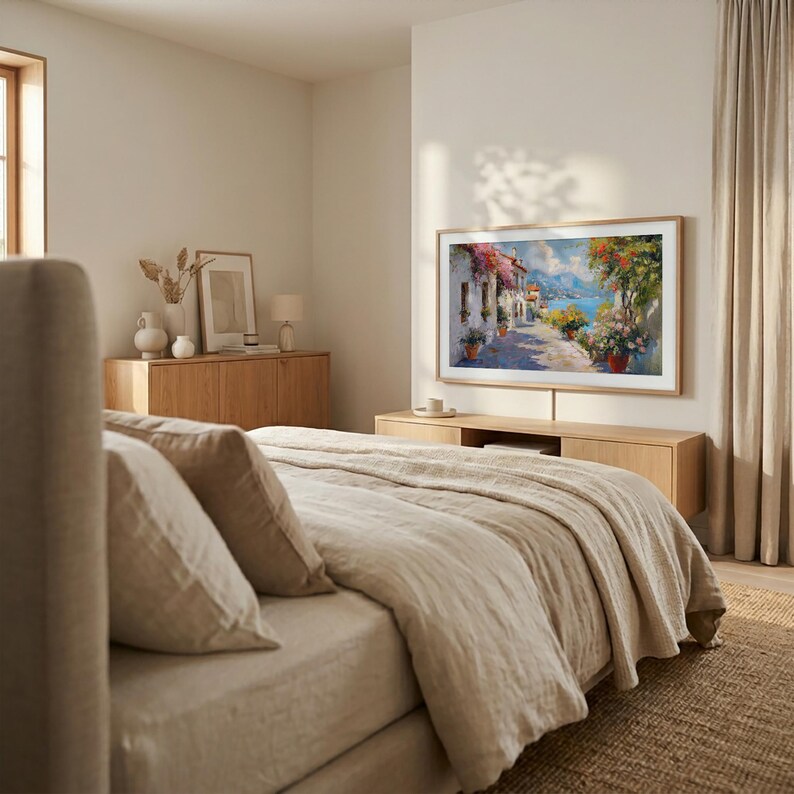 May include: A bedroom with a large framed painting depicting a coastal town. The painting is mounted above a wooden cabinet. A bed with beige bedding and pillows is in the foreground. The room has neutral tones and natural light.