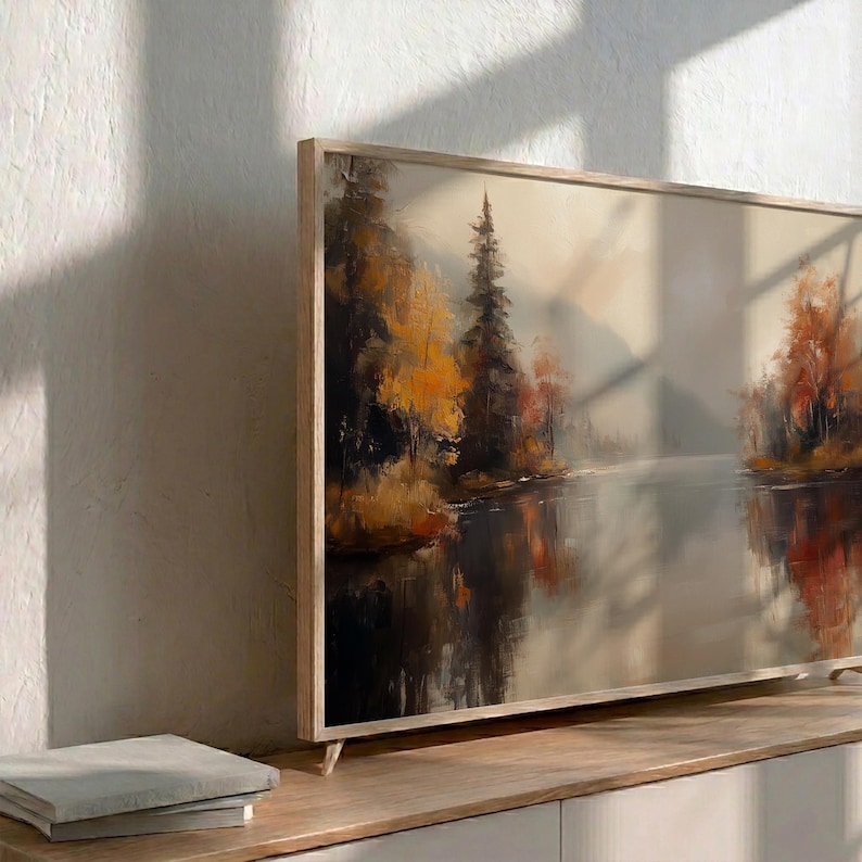 May include: A framed landscape painting depicting a serene lake scene with trees in autumn colors. The artwork features a wooden frame and is displayed on a light-colored wooden surface. The painting's colors include shades of orange, brown, and gray.