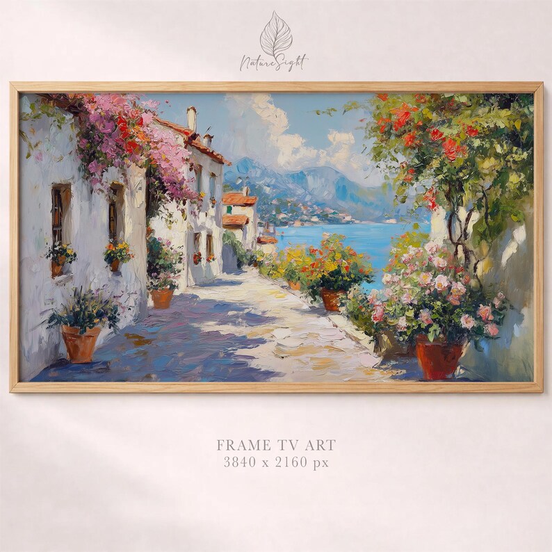 May include: A framed print of a coastal village scene. The artwork shows white buildings with red-tiled roofs, a cobbled street, and colourful flowers. The background features a blue sea and mountains. The frame is made of light-coloured wood.