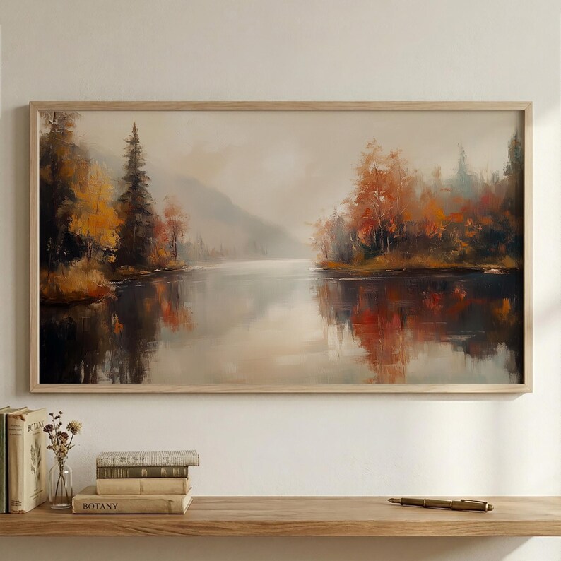 May include: A framed landscape painting depicts a serene lake scene. The artwork features a muted color palette with reflections of autumn trees in shades of orange, yellow, and red. The painting is encased in a light-colored wooden frame, creating a warm and inviting aesthetic.
