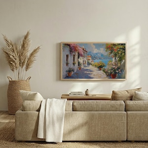 May include: A framed landscape painting shows a sunlit street scene with white buildings, colourful flowers, and a sea view. The artwork is in a light wood frame, displayed above a beige sofa with decorative cushions and a woven basket.
