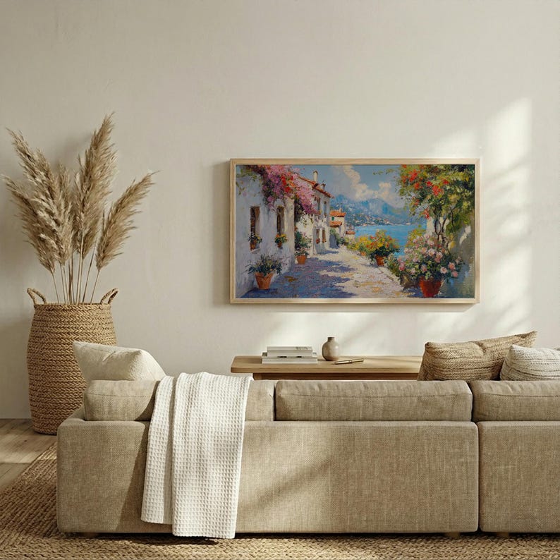 May include: A framed landscape painting shows a sunlit street scene with white buildings, colourful flowers, and a sea view. The artwork is in a light wood frame, displayed above a beige sofa with decorative cushions and a woven basket.