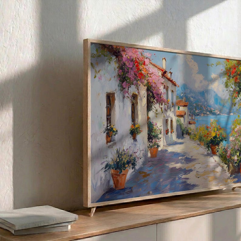 May include: A framed painting of a European village scene, featuring white buildings, a cobbled street, and colourful flowers. The artwork is displayed on a wooden cabinet, with a stack of books in the foreground. The frame is made of light-coloured wood.