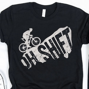 Könnte beinhalten: Schwarzes T-Shirt mit einem weißen Grafikdruck einer Person, die mit dem Fahrrad bergauf fährt, mit dem Text "OH SHIFT" in einer verwitterten Schrift.