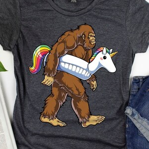 Può includere: Una maglietta grigio scuro con una grafica di un Bigfoot che trasporta un galleggiante per piscina a forma di unicorno. L'unicorno è arcobaleno e il Bigfoot è marrone.