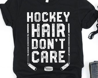 May include: Black t-shirt with white text that reads "Hockey Hair Don't Care". The text is in a distressed font. Two hockey sticks are on either side of the text. A hockey puck is below the text. The t-shirt is laid flat on a white surface. A pair of black trainers and a pair of blue jeans are also on the surface.