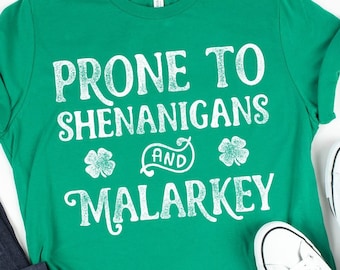 Könnte beinhalten: Grünes T-Shirt mit weißem Text, der "Prone to Shenanigans and Malarkey" lautet. Das Shirt ist gefaltet und liegt auf einer weißen Oberfläche mit einer blauen Jeans, weißen Turnschuhen und einer grünen, kleeblattartigen Brille und Mütze.