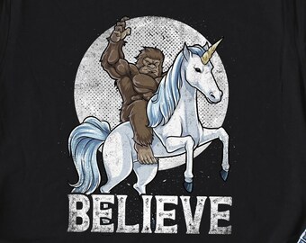 Könnte beinhalten: Schwarzes T-Shirt mit einem Aufdruck eines Bigfoots, der ein Einhorn reitet. Der Aufdruck befindet sich in einem Kreis mit einem Mondhintergrund. Der Text "BELIEVE" befindet sich unter dem Aufdruck.