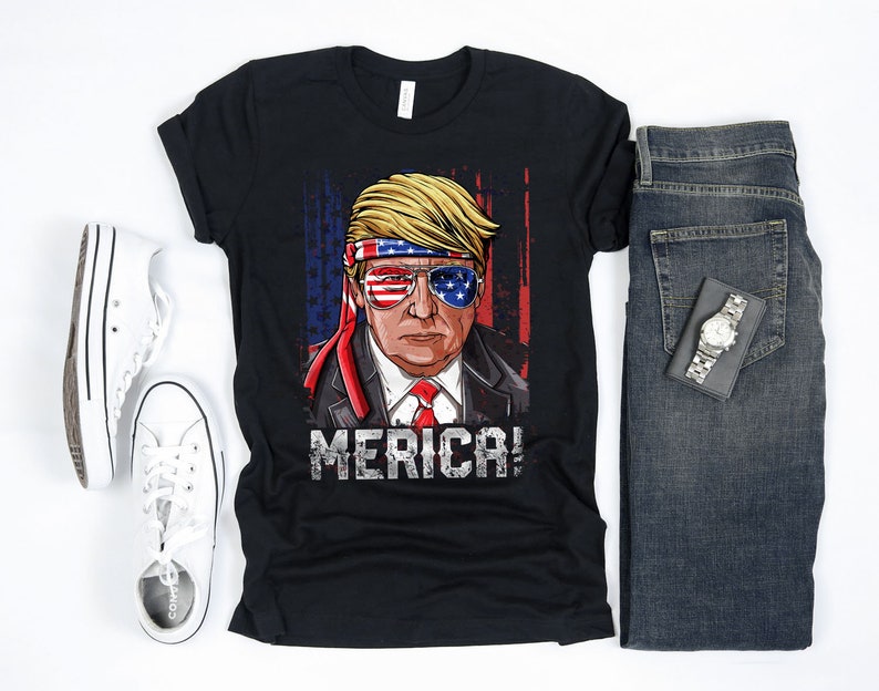 Peut inclure: T-shirt noir avec une image de dessin anim&eacute; d'une personne portant un bandeau et des lunettes de soleil rouge, blanc et bleu. La personne porte une cravate rouge, blanche et bleue et le texte "MERICA!" est imprim&eacute; sous l'image.