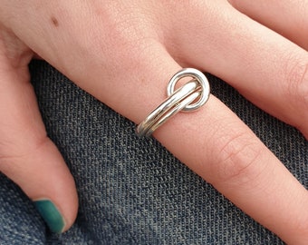 May include: A silver ring with a double knot design. The ring is worn on a finger.