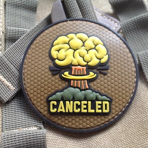 May include: A round patch with a brown background and a black border. The patch features a yellow brain-shaped explosion with orange and green accents. The word "CANCELED" is written in yellow letters at the bottom.