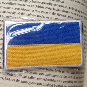 May include: Embroidered patch depicting the Ukrainian flag. The patch is rectangular, with a blue top half and a yellow bottom half, and a white border. The flag is made of fabric.