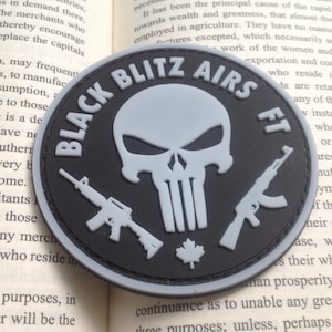 May include: A round, black and gray patch with the text "BLACK BLITZ AIRS FT" encircling a gray skull and two gray rifles. A maple leaf is at the bottom. The patch is on top of an open book.