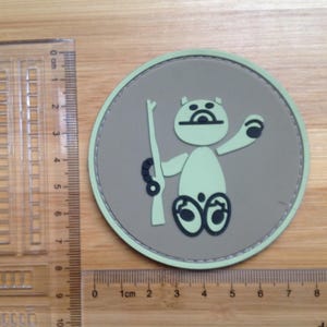 May include: A round patch featuring a cartoon bear holding a rifle. The bear is light green with black details, set against a tan background. The patch has a light green border and is approximately 3 inches in diameter.
