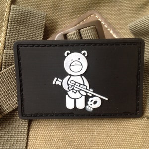 Puede incluir: Parche rectangular negro con un oso de dibujos animados blanco sosteniendo un rifle y una calavera. El parche tiene un borde negro y está sobre un fondo beige.