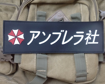 Könnte beinhalten: Ein schwarzer rechteckiger Aufnäher mit weißem japanischem Text und einem rot-weißen Regenschirm-Logo. Der Aufnäher ist auf einem beigefarbenen Rucksack befestigt. Der japanische Text lautet "アンブレラ社".