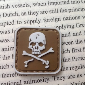 May include: A brown and white embroidered patch with a white skull and crossbones design. The patch has a stitched border.