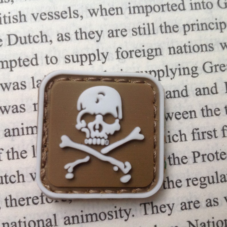 May include: A brown and white embroidered patch with a white skull and crossbones design. The patch has a stitched border.
