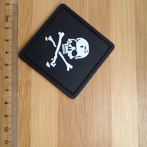 May include: A black diamond-shaped patch with a white skull and crossbones design. The patch is approximately 5 cm wide.