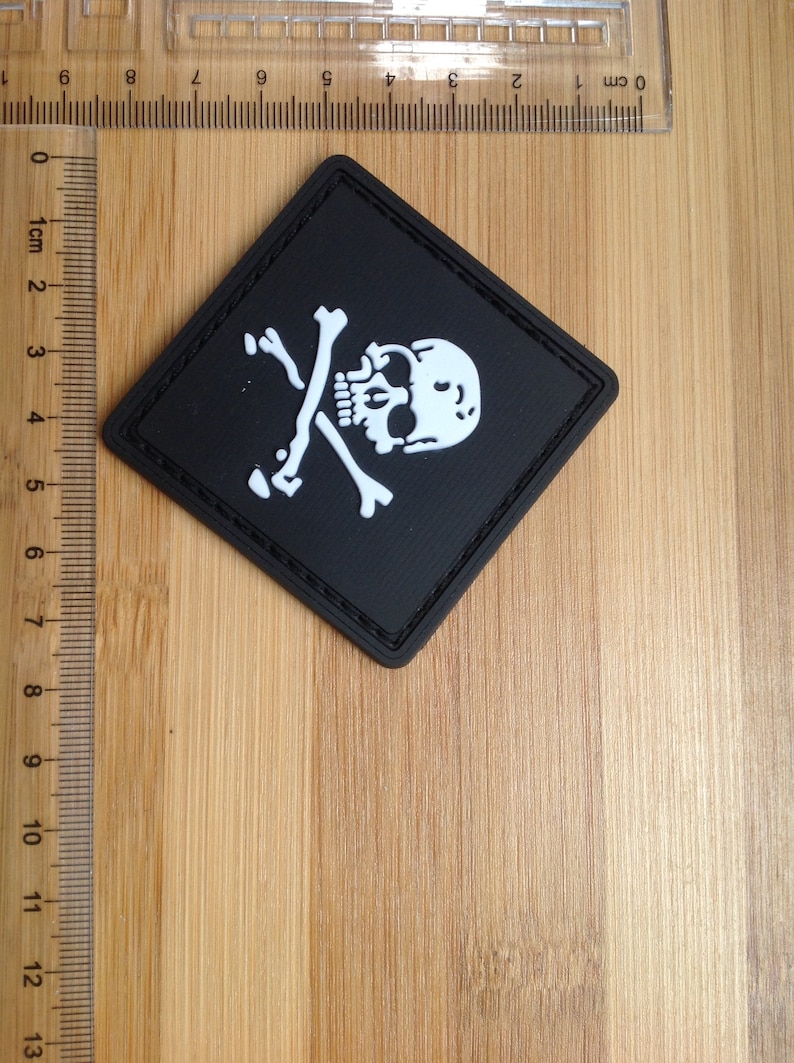 May include: A black diamond-shaped patch with a white skull and crossbones design. The patch is approximately 5 cm wide.
