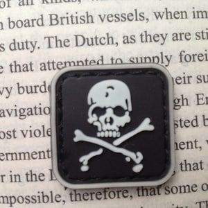 May include: A black and white embroidered patch with a skull and crossbones design. The skull is white with black eye sockets and a black nose. The crossbones are white and are crossed behind the skull.