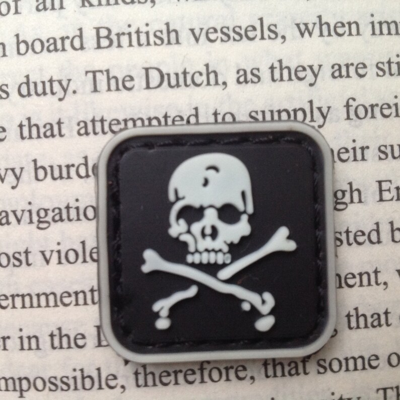 May include: A black and white embroidered patch with a skull and crossbones design. The skull is white with black eye sockets and a black nose. The crossbones are white and are crossed behind the skull.