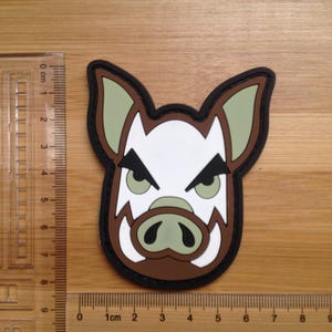 Puede incluir: Un parche con la cabeza estilizada de un jabalí con contorno negro. El diseño incluye un hocico blanco, colmillos marrones y orejas verdes. El parche mide aproximadamente 7.6 cm de ancho.