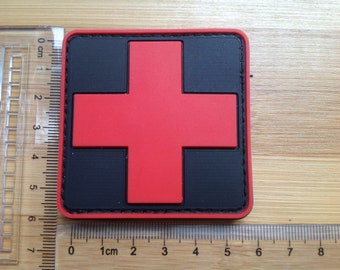 May include: A square patch featuring a red cross on a black background, with a black border. The patch is approximately 2.5 inches wide and 2.5 inches tall. The patch is on a wooden surface.