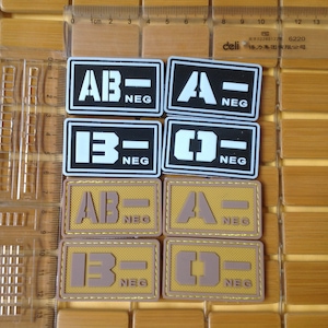 May include: Eight black and tan fabric patches with white or tan lettering. The patches are rectangular and have a stitched border. The patches display the blood types AB-, A-, B-, and O- with the word "NEG" below each blood type.