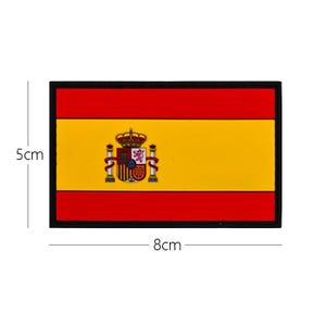 Puede incluir: Parche rectangular con el diseño de la bandera española. El parche tiene una franja roja superior e inferior, un centro amarillo y un borde negro. El escudo de armas español está en el centro. Mide aproximadamente 8 cm de ancho y 5 cm de alto.
