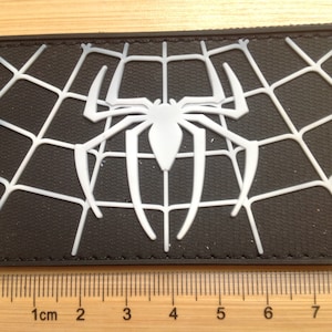 May include: A rectangular black patch with a white spider and web design. The spider is central, with its legs and body extending outwards. The web is intricately designed. The patch is approximately 7.6 cm wide.
