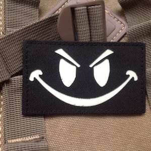 May include: A black patch with a white smiling face with angry eyes. The patch is sewn onto a brown fabric background.